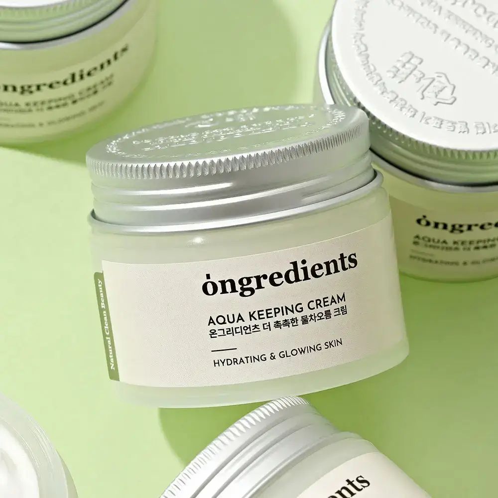 Mehrere Creme-Gläser mit silbernen Deckeln. Aufschrift: Ongredients, Aqua Keeping Cream, Hydrating & Glowing Skin. Auf grünem Hintergrund.