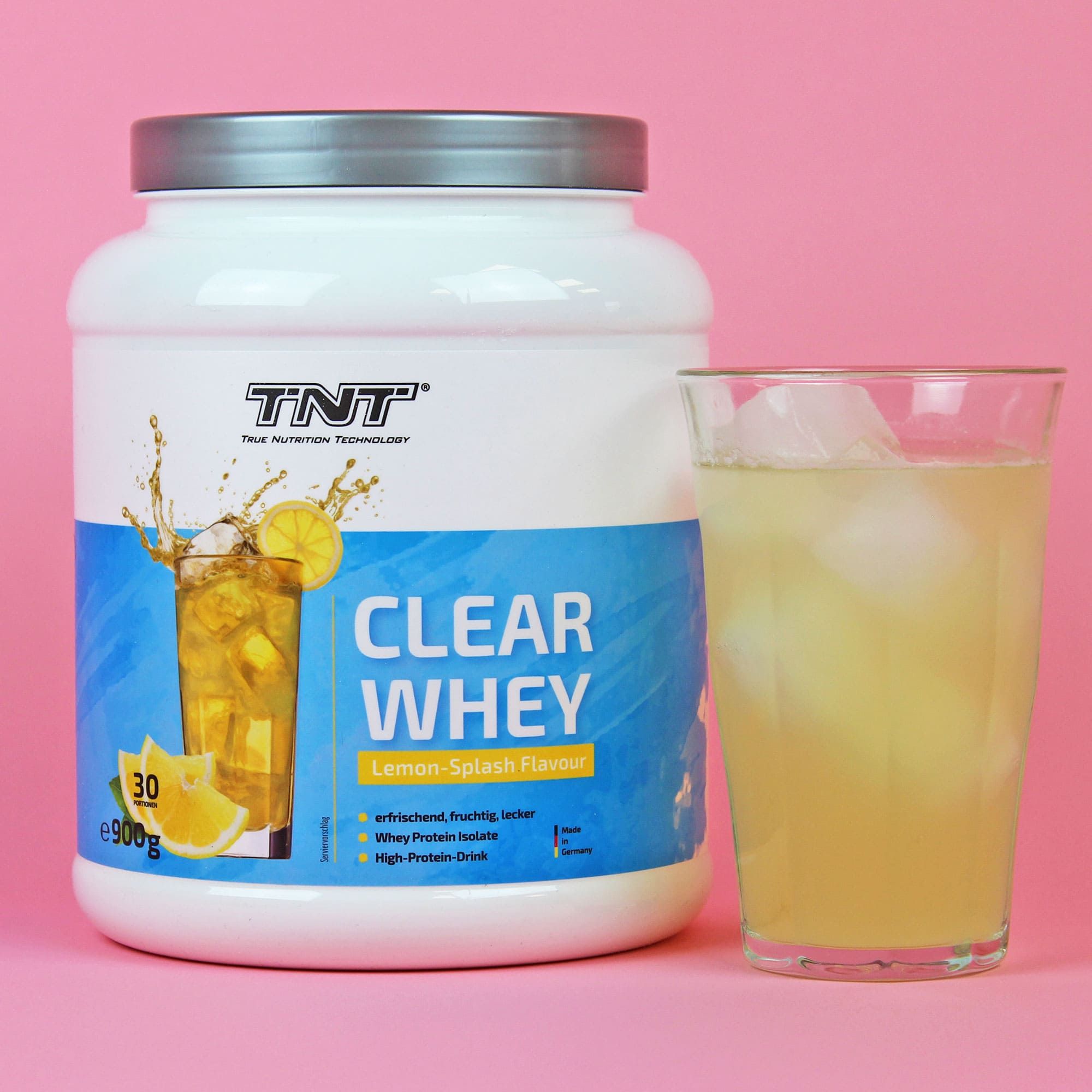 TNT CLEAR WHEY-Behälter und Glas mit Getränk. Aufdruck: CLEAR WHEY, Lemon Splash Flavour. Hintergrund: Pink.