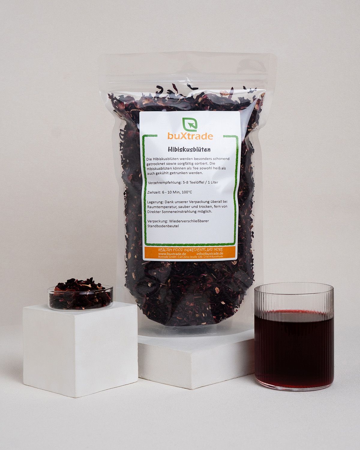 Beutel mit Hibiskusblüten, Glas mit Getränk, Schale mit Blüten. Auf weißem Podest. Produktetikett sichtbar.