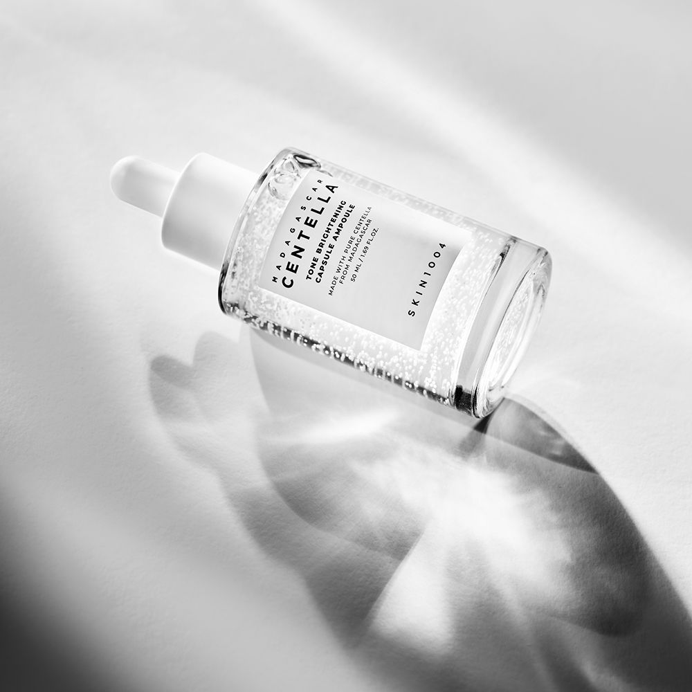 Schräg liegende Glasflasche mit weißem Deckel. Enthält Flüssigkeit mit kleinen Partikeln. Text: MADAGASCAR CENTELLA, Tone Brightening Capsule Ampoule.