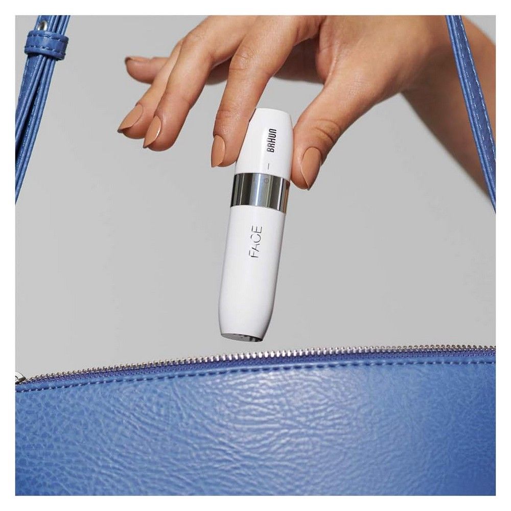Hand hält einen weißen Braun Gesichts-Haarentferner über einer blauen Handtasche.