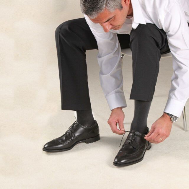 Mann bindet Schnürsenkel. Er trägt schwarze Kompressionsstrümpfe, schwarze Schuhe, eine weiße Hemd und eine schwarze Hose.