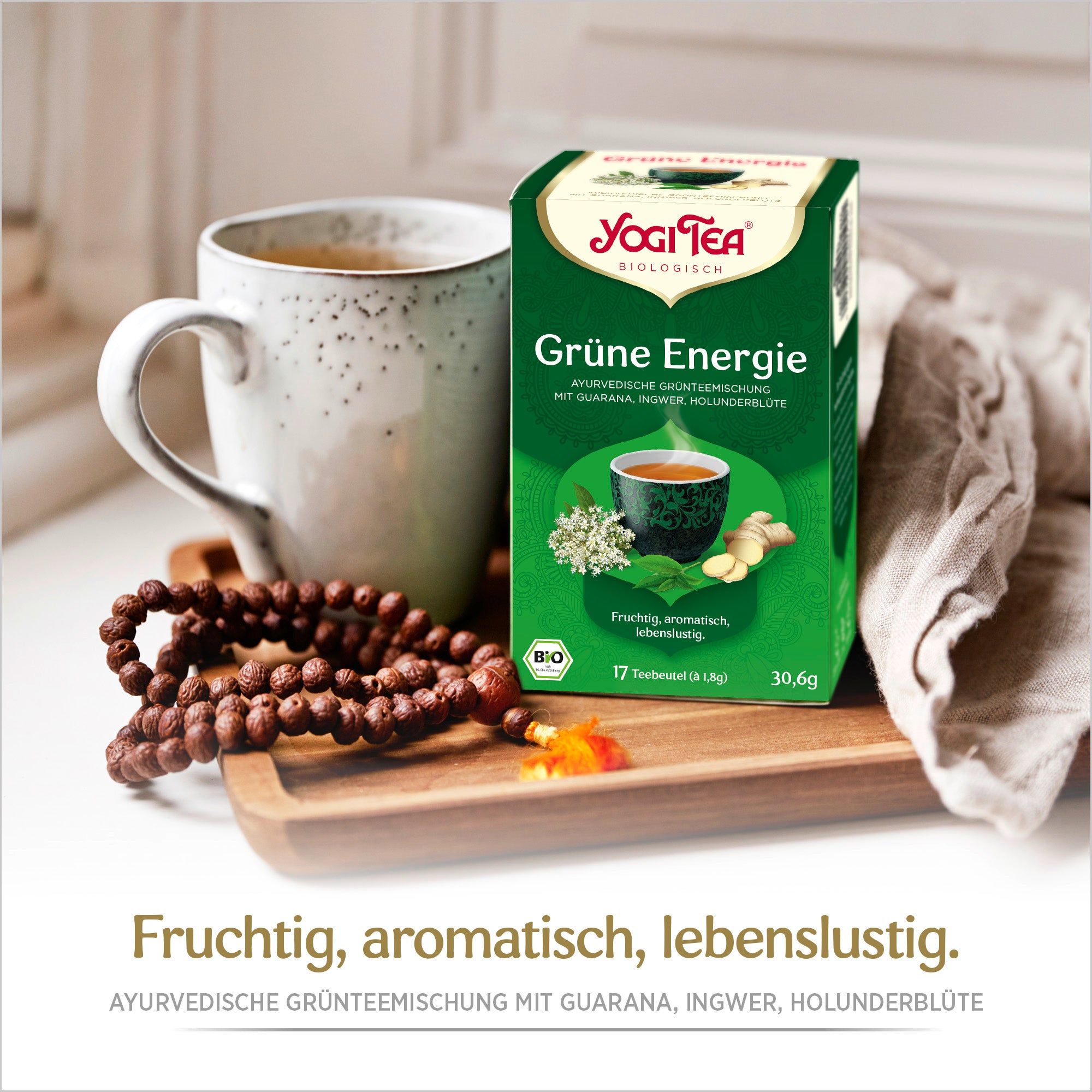 Yogi Tea-Schachtel neben Tasse Tee. Aufschrift: Grüne Energie, ayurvedische Grünteemischung. Bio-Siegel. 17 Teebeutel. Holztablett, Kette.