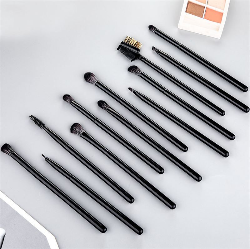 Zwölf schwarze Lidschatten- und Augenbrauenpinsel, auf einer grauen Oberfläche liegend, mit einem Lidschatten-Set im Hintergrund.