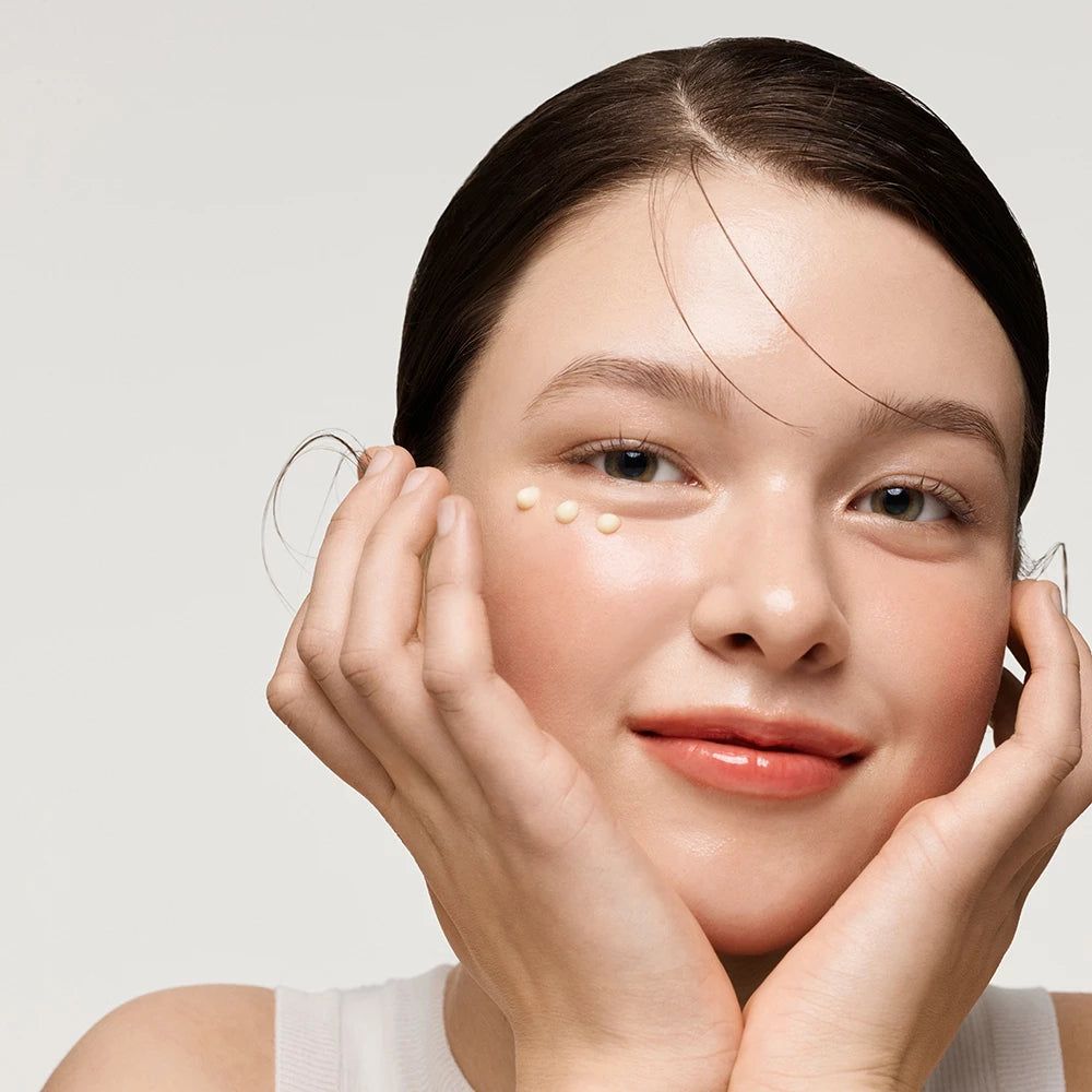 Porträt einer Frau mit Creme-Punkten unter den Augen. Sie lächelt. Hände am Gesicht.