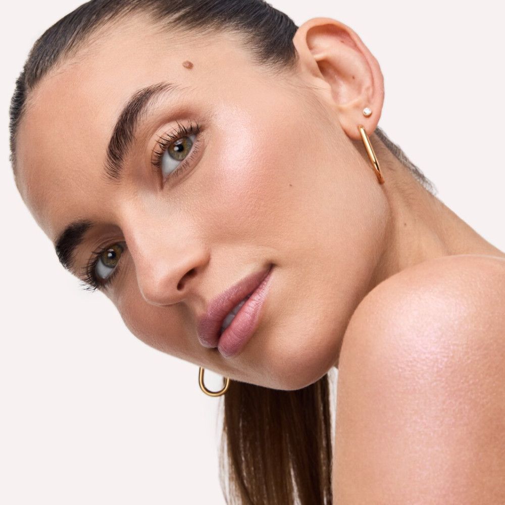 Nahaufnahme einer Frau mit Highlighter auf der Haut. Sie trägt goldene Ohrringe. Ihr Blick geht zur Kamera.