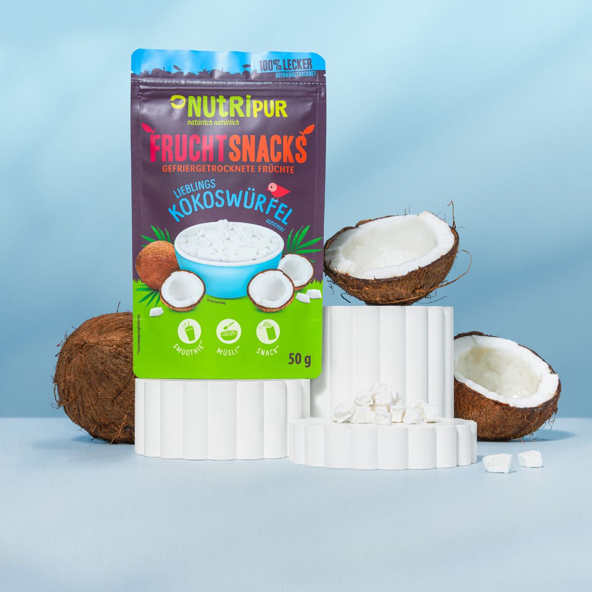 NutriPur Frucht Snacks Kokoswürfel Verpackung. Kokosnuss-Hälften und Würfel auf Podesten. 50g Packung.