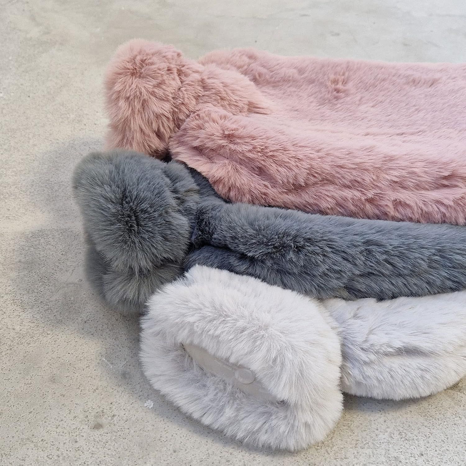 Drei Wärmflaschen mit flauschigen Bezügen. Rosa, grau und weiß. Rechteckige Form. Weiches Material. Teilweise sichtbar.