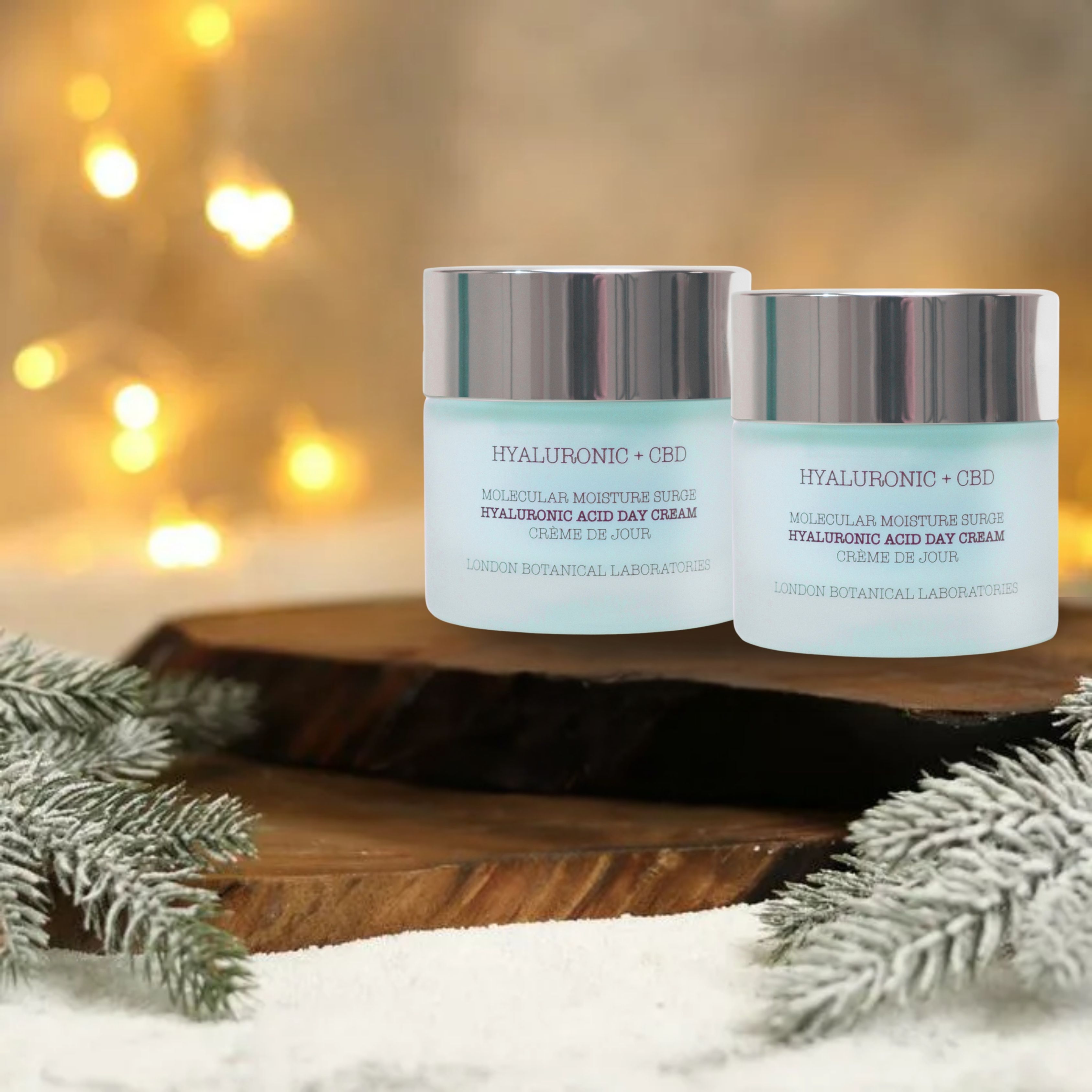 Zwei Tiegel Tagescreme mit Text: Hyaluronsäure + CBD. Auf Holzbrett, mit Tannenzweigen und Bokeh-Effekt im Hintergrund.