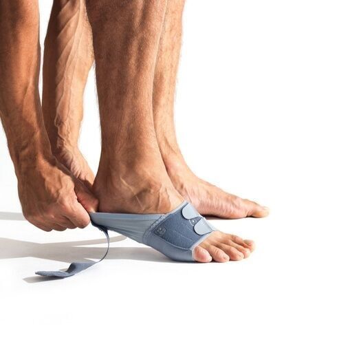 Person zieht an einem Teil der grau-blauen Fußbandage. Klettverschluss und gepolsterte Bereiche sind sichtbar. Der Fuß ist teilweise bedeckt.