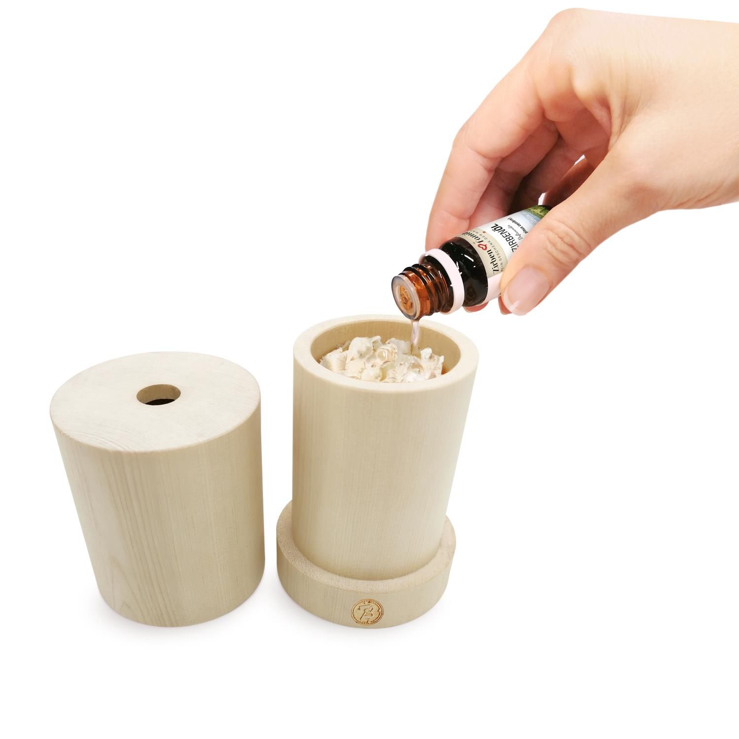 Hand gießt Zirbenöl aus einer Flasche in einen Holzdiffusor. Daneben liegt der Deckel des Diffusors.