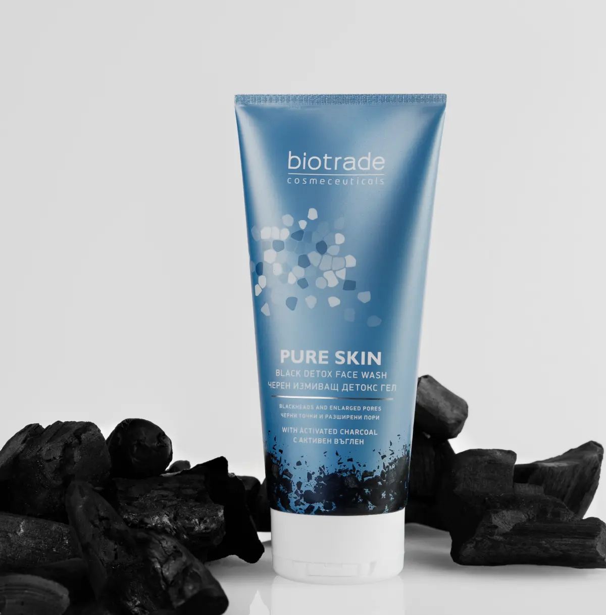 Blau-graue Tube mit weißem Deckel, umgeben von Holzkohle. Aufschrift: Biotrade, Pure Skin Black Detox Face Wash.