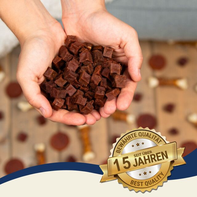 Hände halten eine Menge brauner, würfelförmiger Snacks. Aufkleber mit der Aufschrift 'BEWAHRTE QUALITÄT SEIT 15 JAHREN BEST QUALITY'.