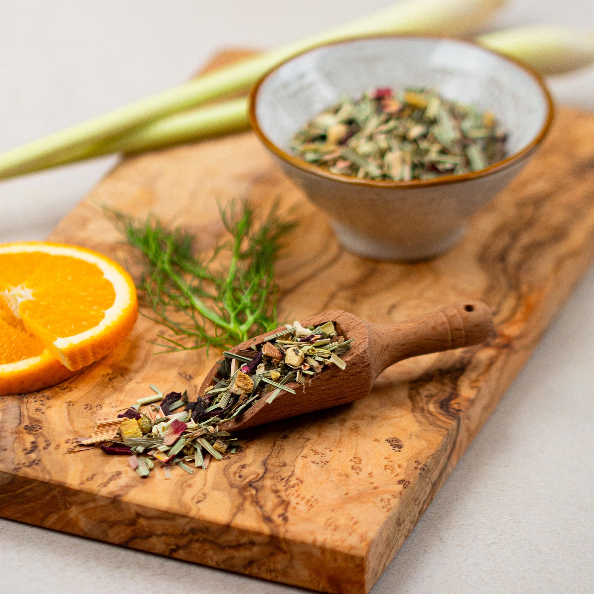 Holzbrett mit Orangenscheibe, Fenchel, Holzlöffel mit Kräutern, Schale mit Kräutern. Hintergrund: Zitronengras.