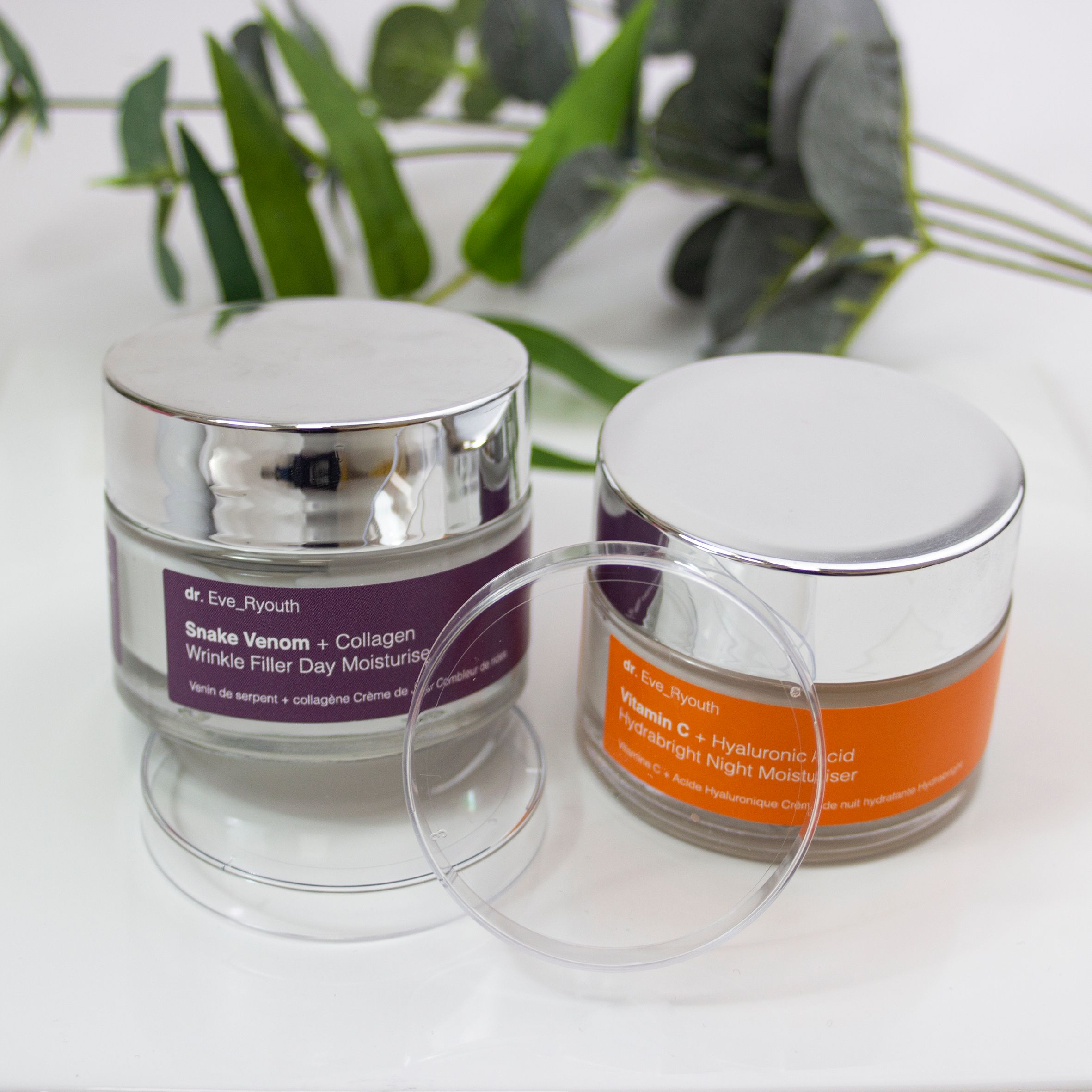 Zwei Tiegel Dr. Eve_Ryouth Tages- und Nachtcreme auf weißem Teller. Violette und orangefarbene Etiketten. Silberne Deckel.