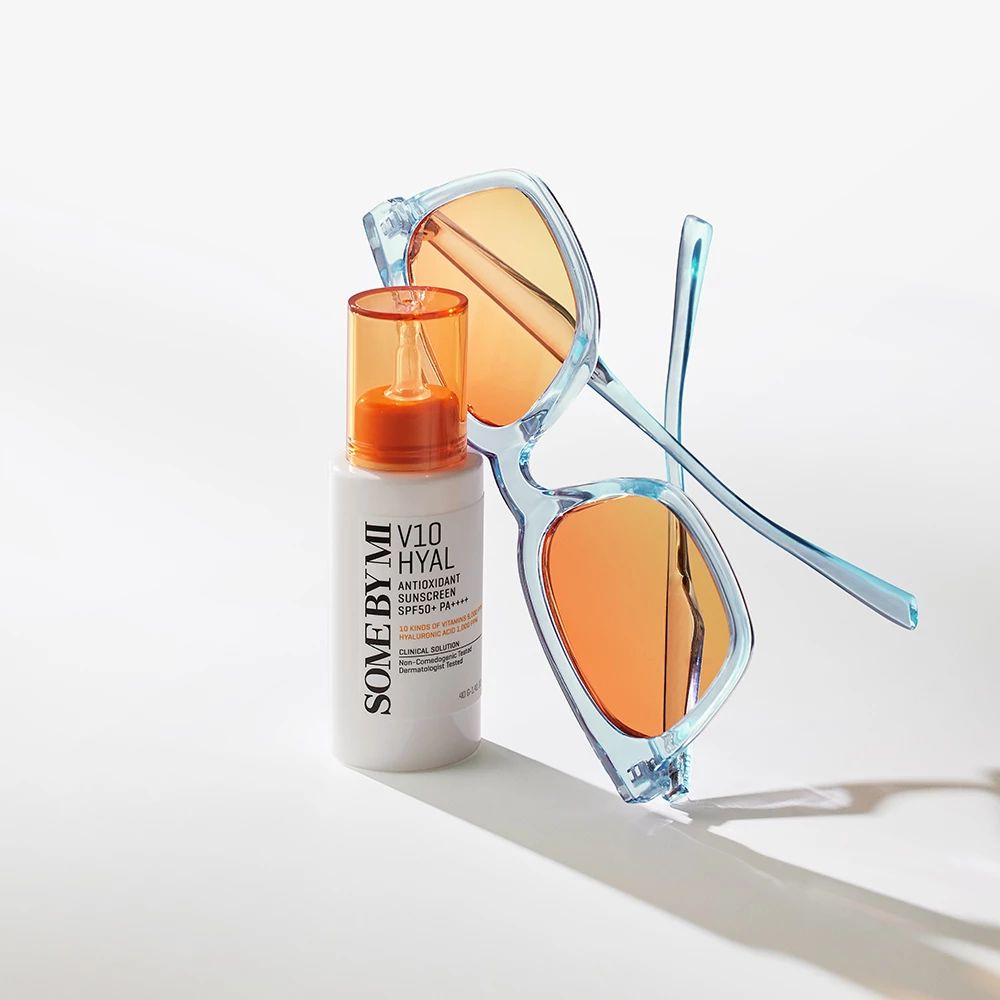Weißer Flakon mit orangefarbenem Deckel neben blauer Sonnenbrille. Text: V10 HYAL Antioxidant Sunscreen SPF50+.