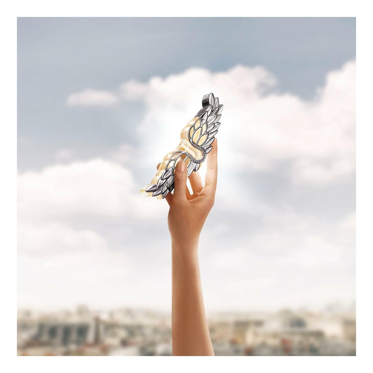 Hand hält silberfarbenes Flügel-Emblem vor Himmel mit Stadt im Hintergrund. Emblem-Design, gehalten.