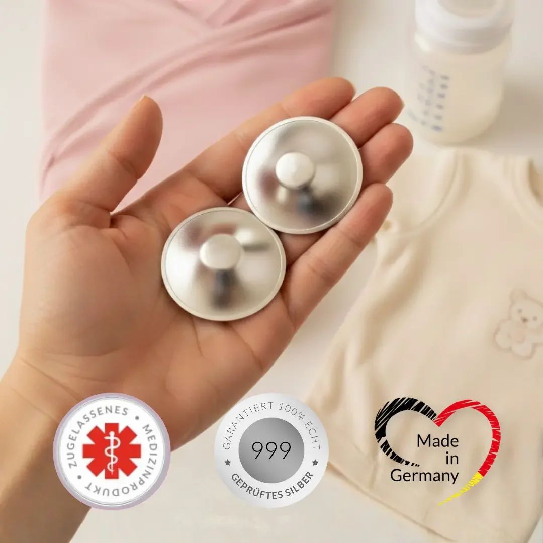 Zwei silberfarbene Brustwarzenhütchen in einer Hand. Medizinprodukt-Siegel, 999 Silber-Siegel, Made in Germany-Siegel.