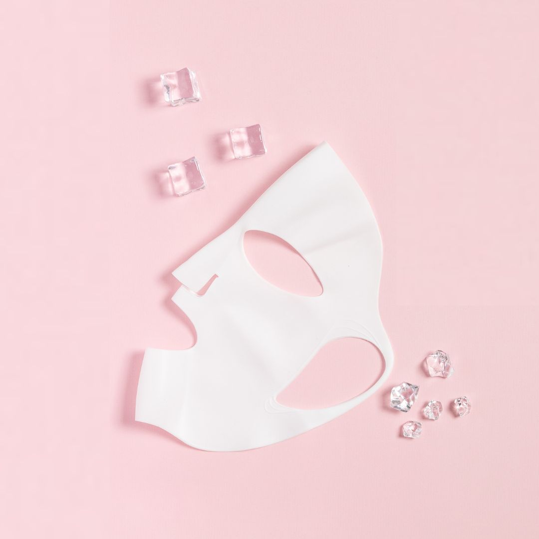 Weiße Silikonmaske auf rosa Hintergrund. Eiswürfel liegen daneben. Maske hat Aussparungen für Augen und Mund.