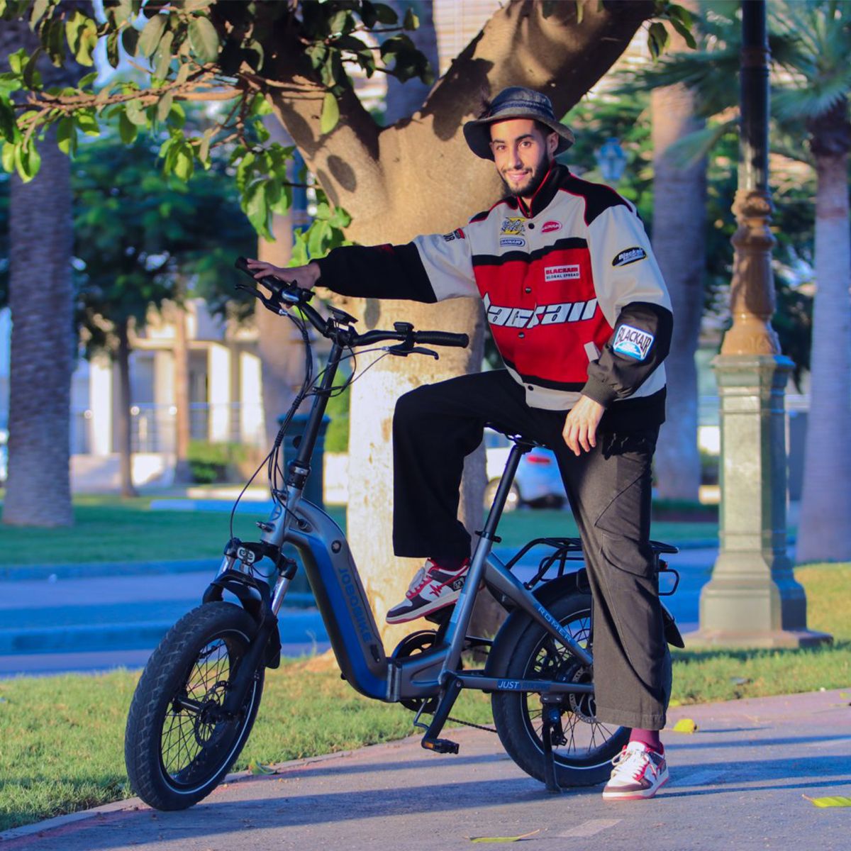 Person sitzt auf einem Elektrofahrrad. Der Mann trägt eine Jacke und einen Hut. Das Fahrrad ist grau und blau. Hintergrund ist eine Straße.