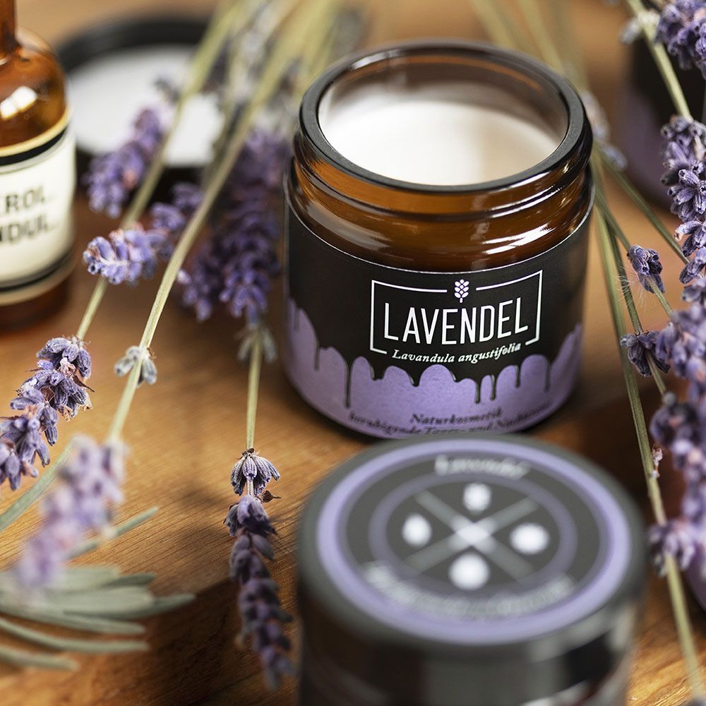Braunes Glas mit weißer Creme, schwarzem Etikett mit Lavendel-Schriftzug. Lavendelzweige umgeben das Produkt.
