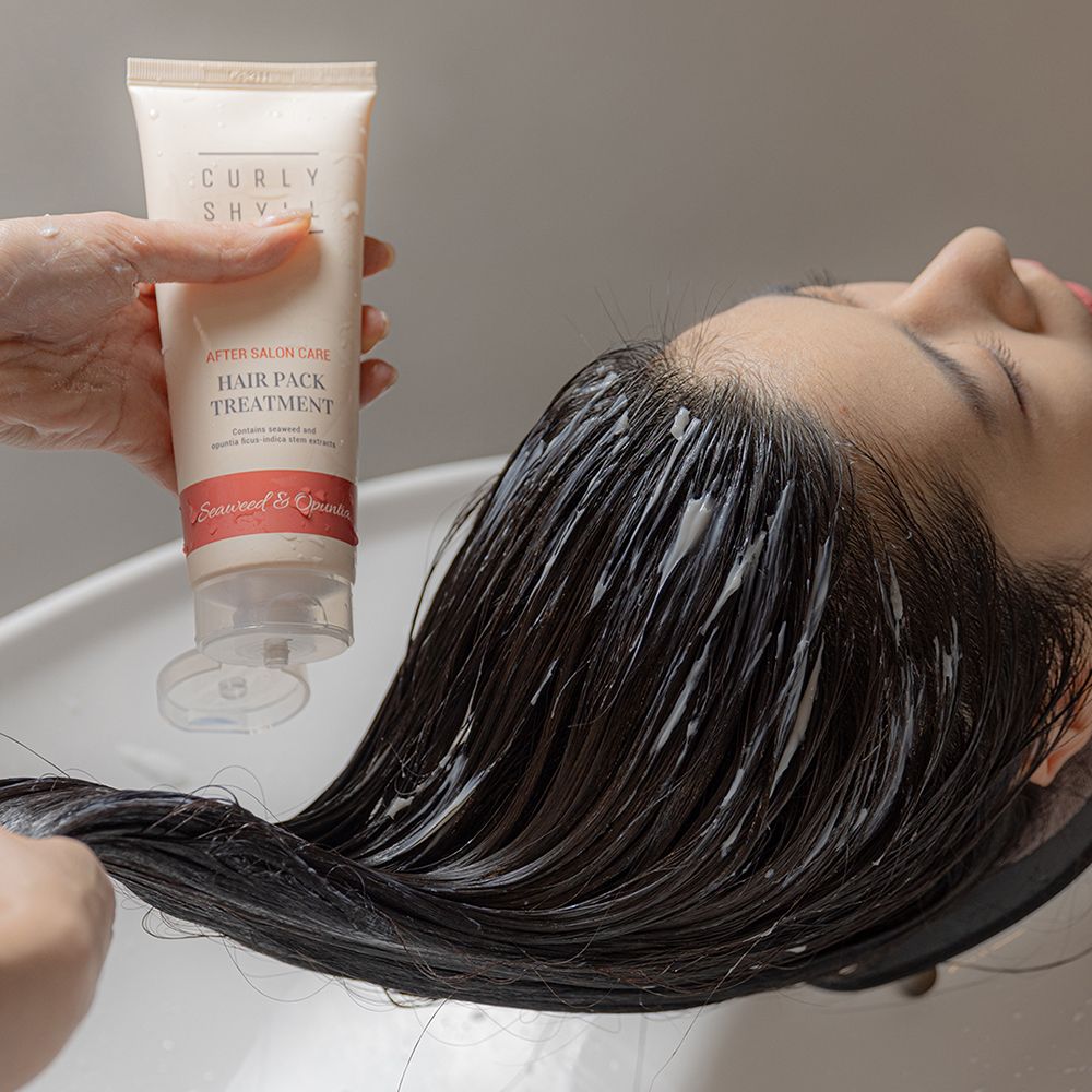 Person hält Tube über Haaren. Aufschrift: CURLY SHYLL, After Salon Care, Hair Pack Treatment. Roter Streifen mit Text: Seaweed & Opuntia.