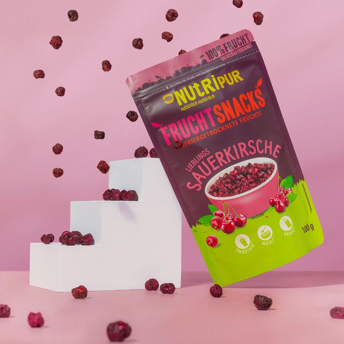 Beutel mit gefriergetrockneten Sauerkirschen, umgeben von Kirschen. Rosa Hintergrund. Aufschrift: NutriPur, Frucht Snacks, 100g.