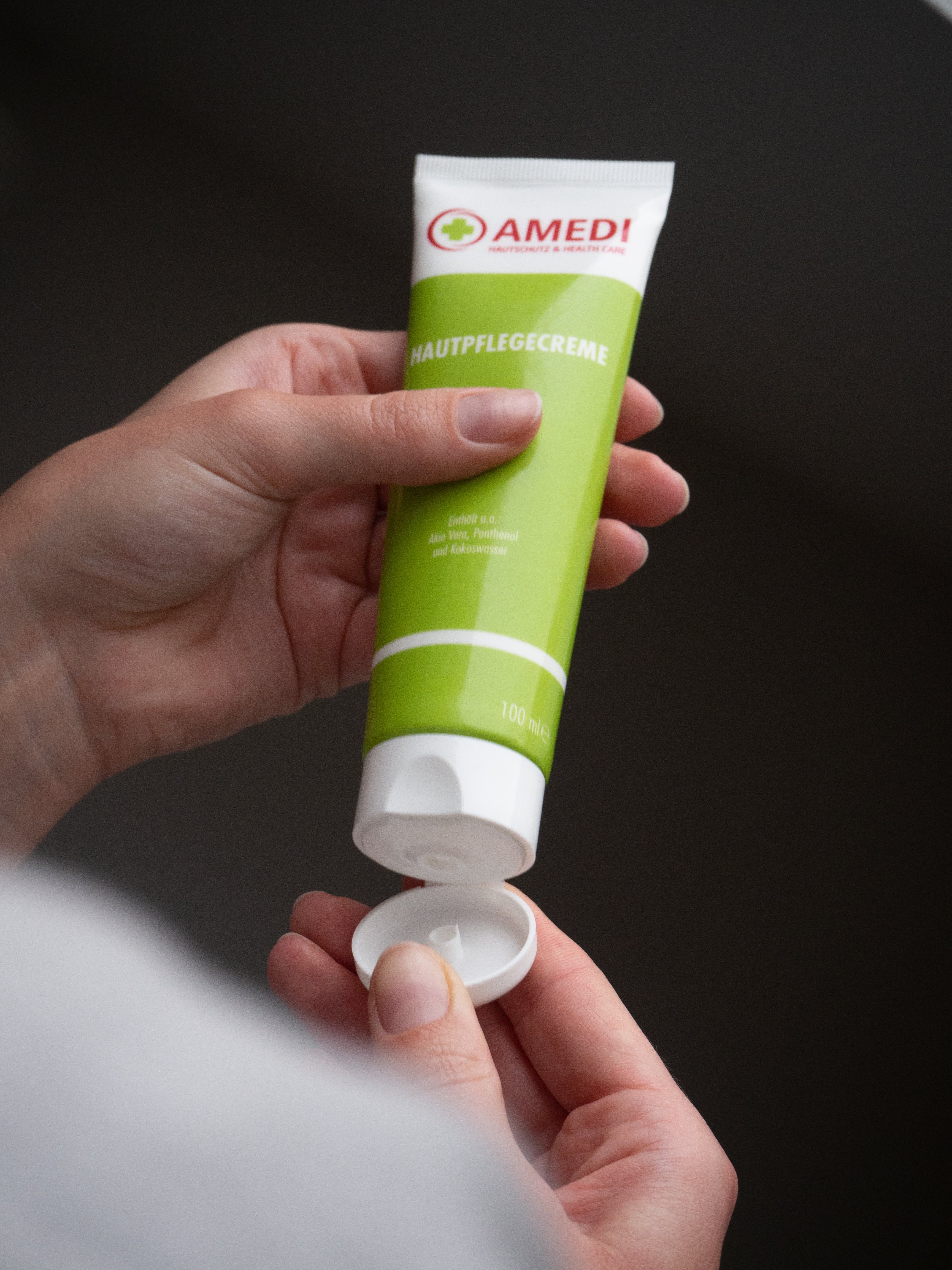 Hand öffnet grüne Tube mit weißem Deckel. Aufschrift: AMEDI, Hautpflegecreme. Logo mit rotem Kreuz. Text in Deutsch.
