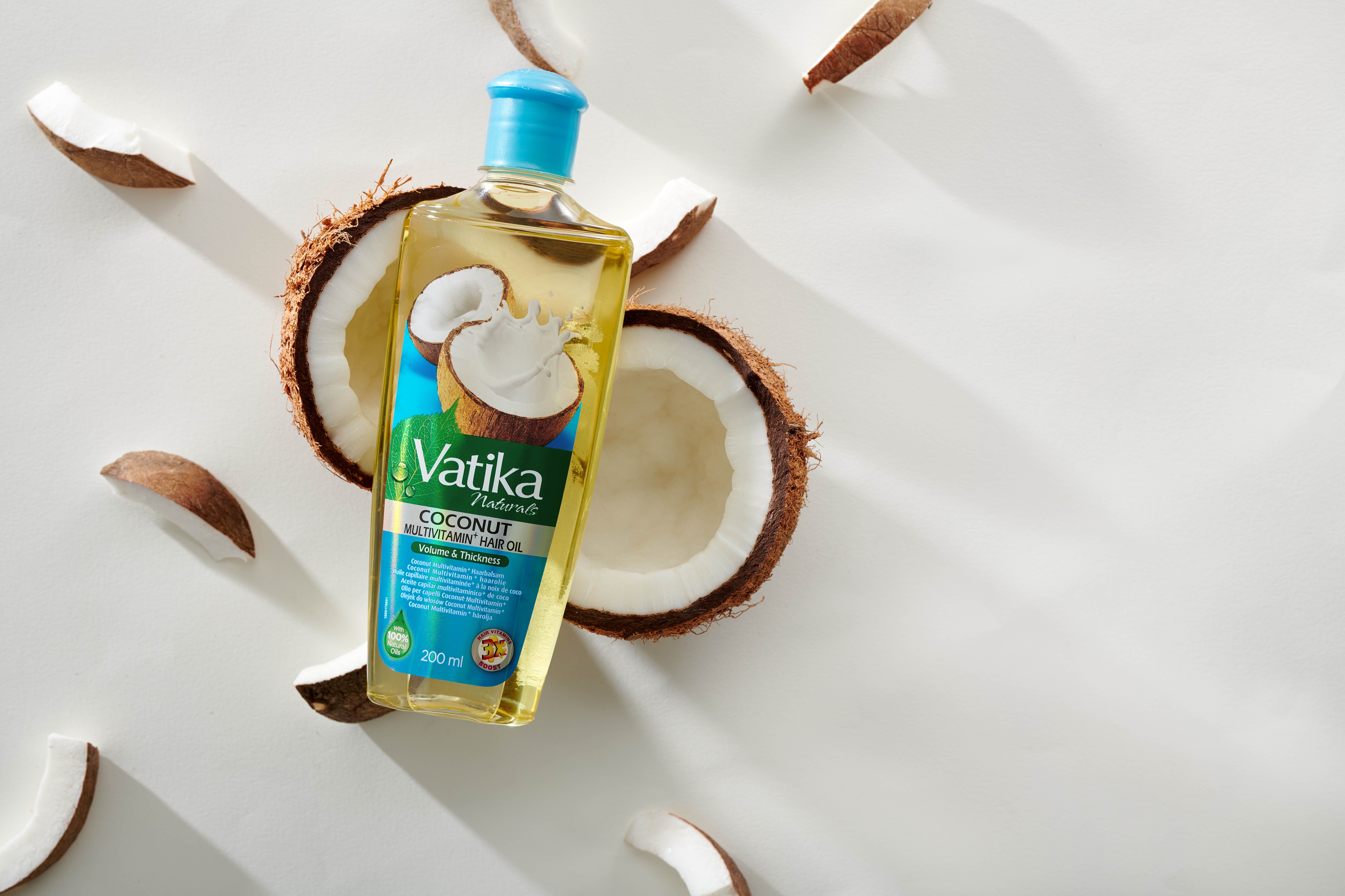 Vatika Naturals Haaröl-Flasche mit blauem Deckel, umgeben von Kokosnuss-Hälften und -Stücken. Aufschrift: Coconut Multivitamin Hair Oil.