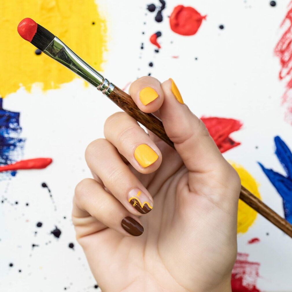 Hand mit gelb und braun lackierten Nägeln, hält Pinsel mit roter Farbe. Hintergrund mit Farbspritzern.