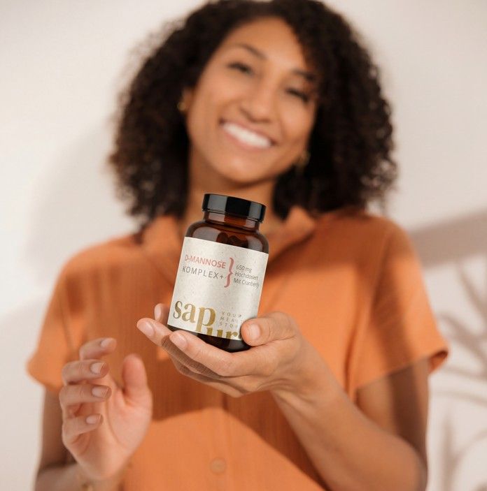 Person hält eine braune Flasche mit Etikett. Aufschrift: D-Mannose Komplex+, 650mg, Mit Cranberry, sapura health.