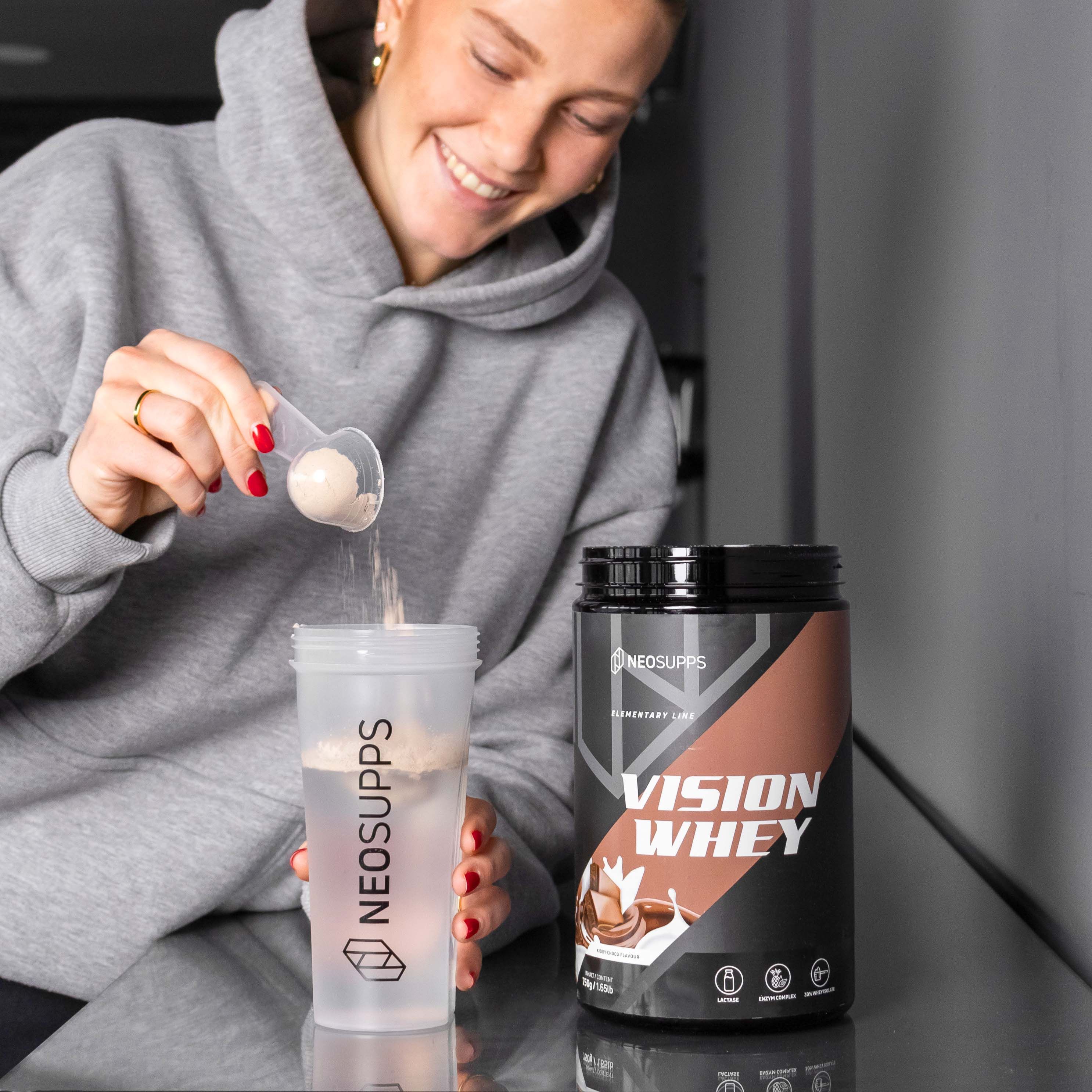 Frau gießt Pulver in Shaker. Dose Vision Whey. Aufschrift: Vision Whey, Kiddy Choco. Braune Dose mit schwarzem Deckel. Shaker.
