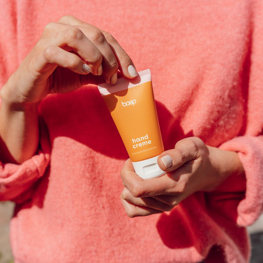 Orange Tube mit rosa Deckel. Aufschrift: boep, hand creme. Hände halten die Tube.