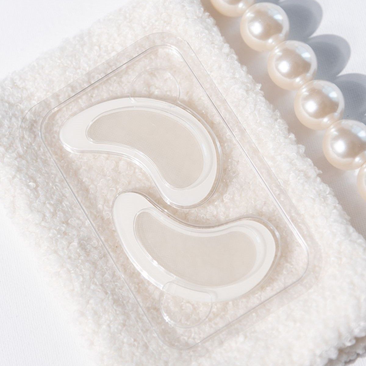 Zwei Augenpads in einer transparenten Verpackung. Auf einem weißen Handtuch mit Perlen.