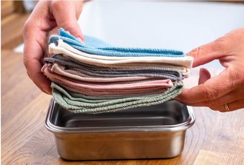 Hände halten einen Stapel farbiger Tücher über einem Behälter. Tücher in verschiedenen Farben. Behälter aus Metall.