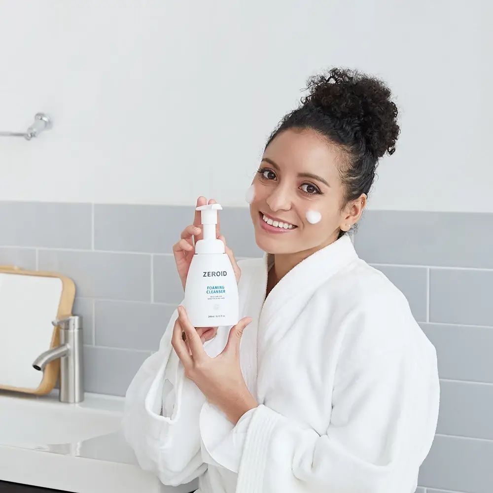 Frau hält eine weiße ZEROID Schaumreiniger-Flasche. Text: FOAMING CLEANSER, mild care for sensitive & dry skin. 240ml/8.12 fl oz.