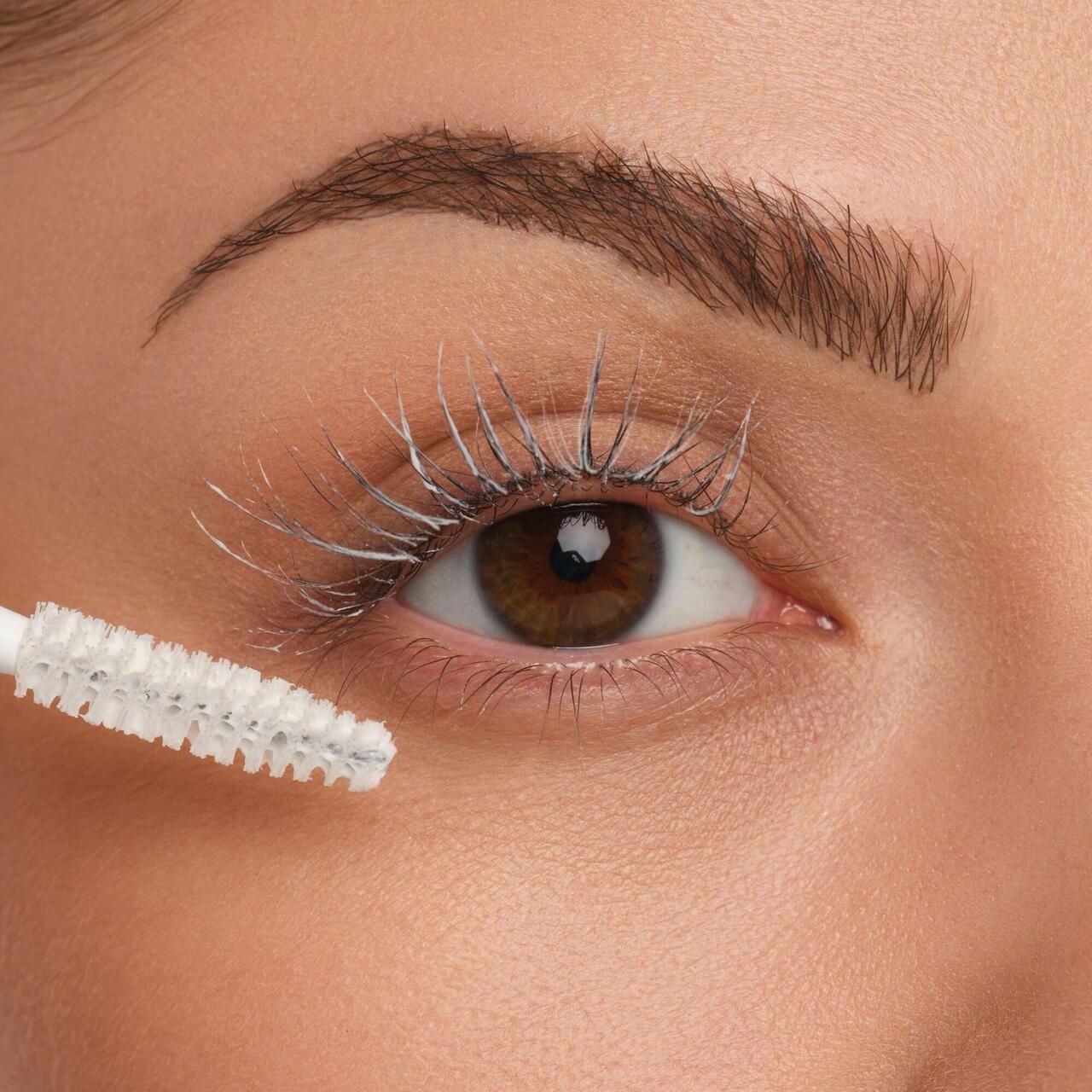 Auge mit Wimpern, die mit weißer Mascara-Basis bedeckt sind. Bürste in der Nähe des Auges.