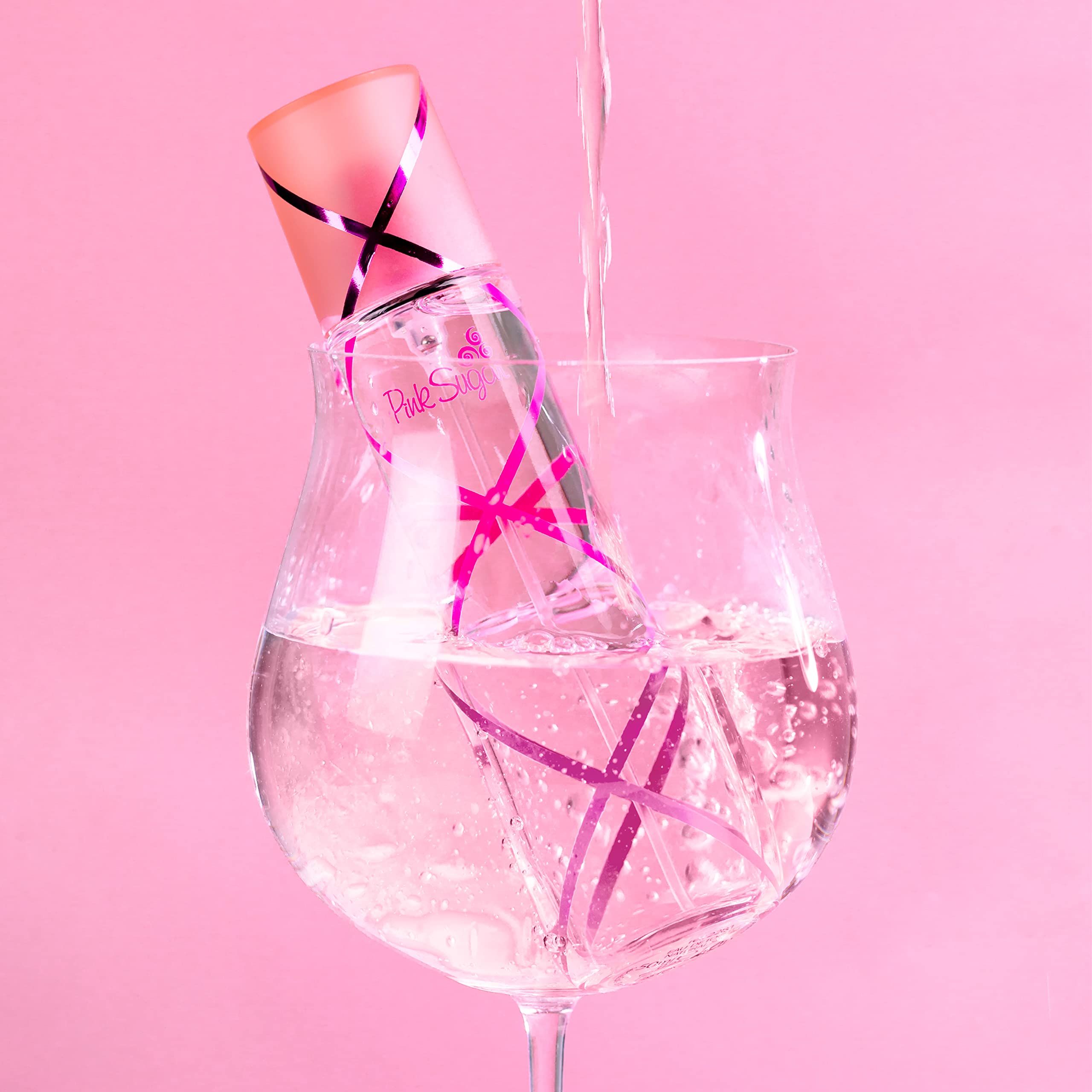 Parfümflasche in einem Glas Wasser. Rosa Akzente und Schriftzug Pink Sugar. Wasser wird in das Glas gegossen.