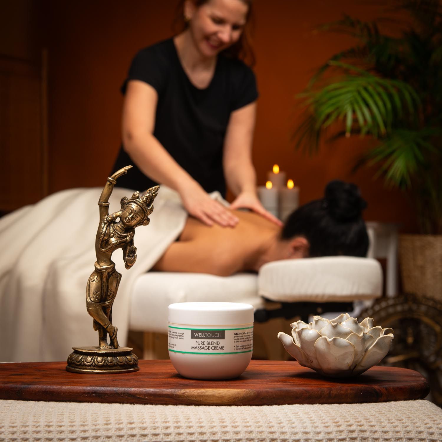 Weißes Creme-Gefäß mit grüner Beschriftung. Aufschrift: Welltouch Pure Blend Massage Creme. Auf Holztablett, vor Massage-Szene.