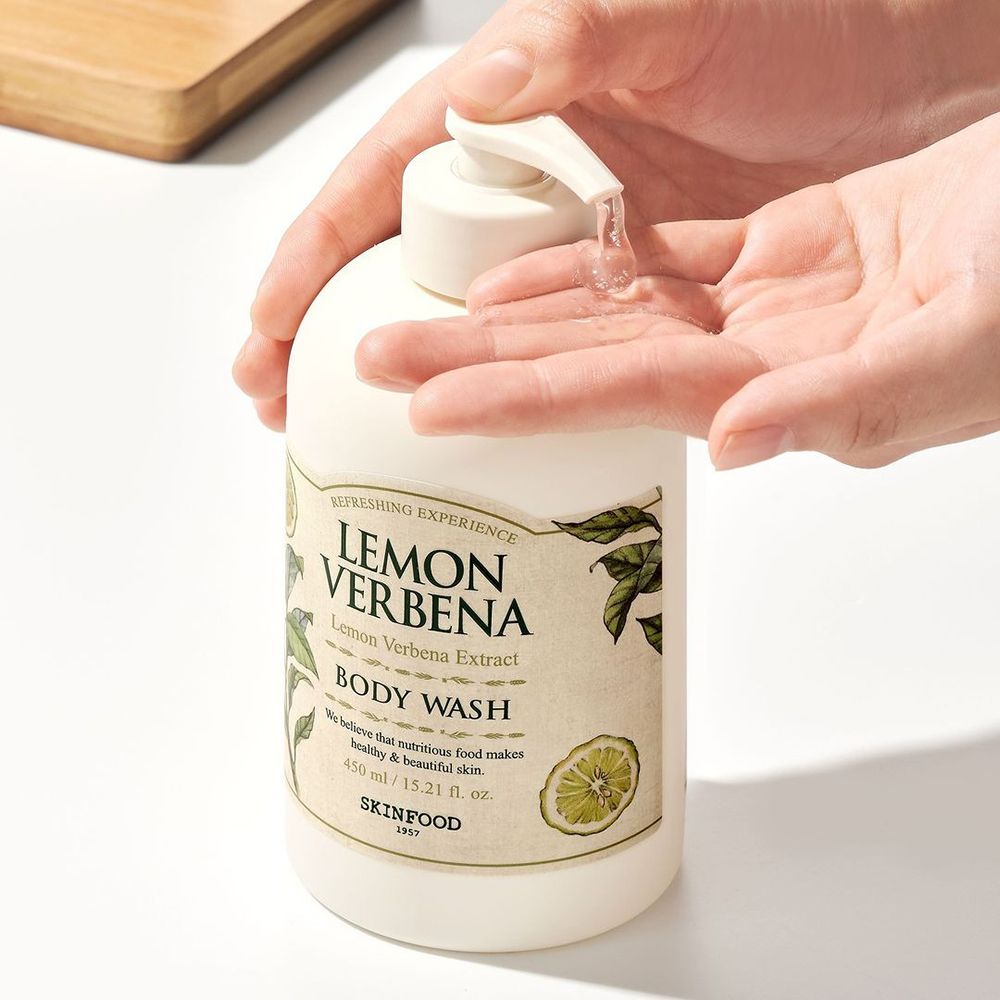 Hände halten SKINFOOD Lemon Verbena Body Wash Flasche. Klare Flüssigkeit wird auf die Hand gepumpt. Produktetikett sichtbar.