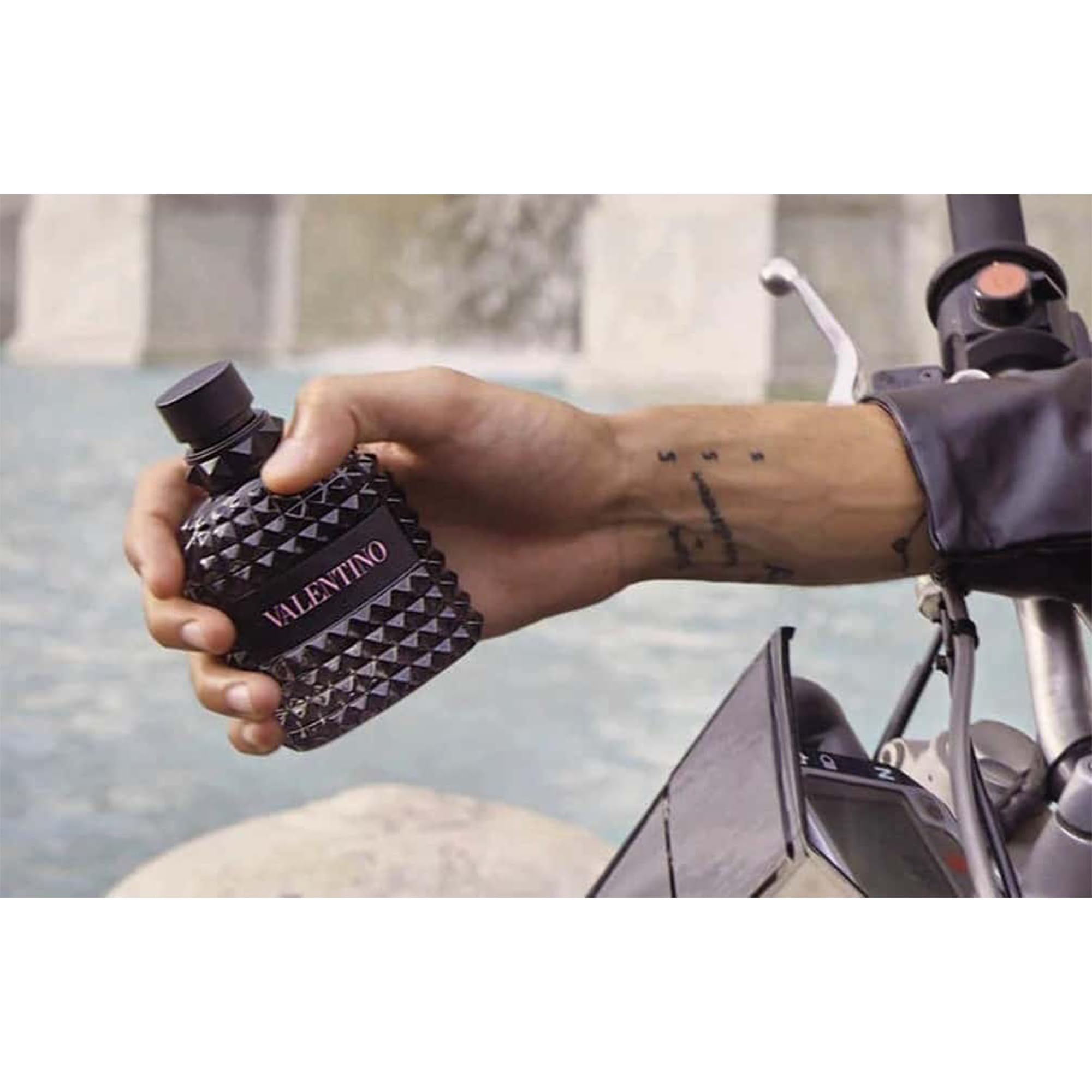 Valentino-Flakon in Hand gehalten. Tattoo am Arm. Hintergrund: Motorrad und Wasser.