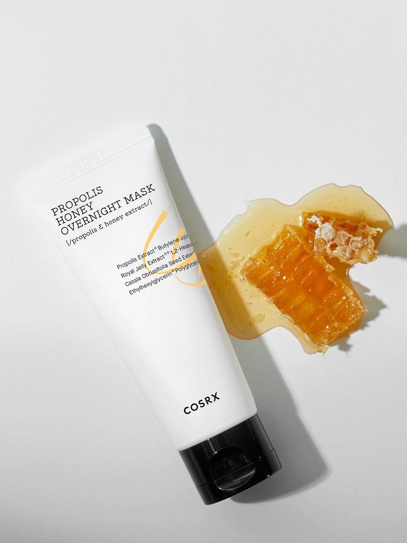 Weiße Tube mit schwarzem Deckel neben Honigwabe und Honig. Aufschrift: PROPOLIS HONEY OVERNIGHT MASK. Marke: COSRX.