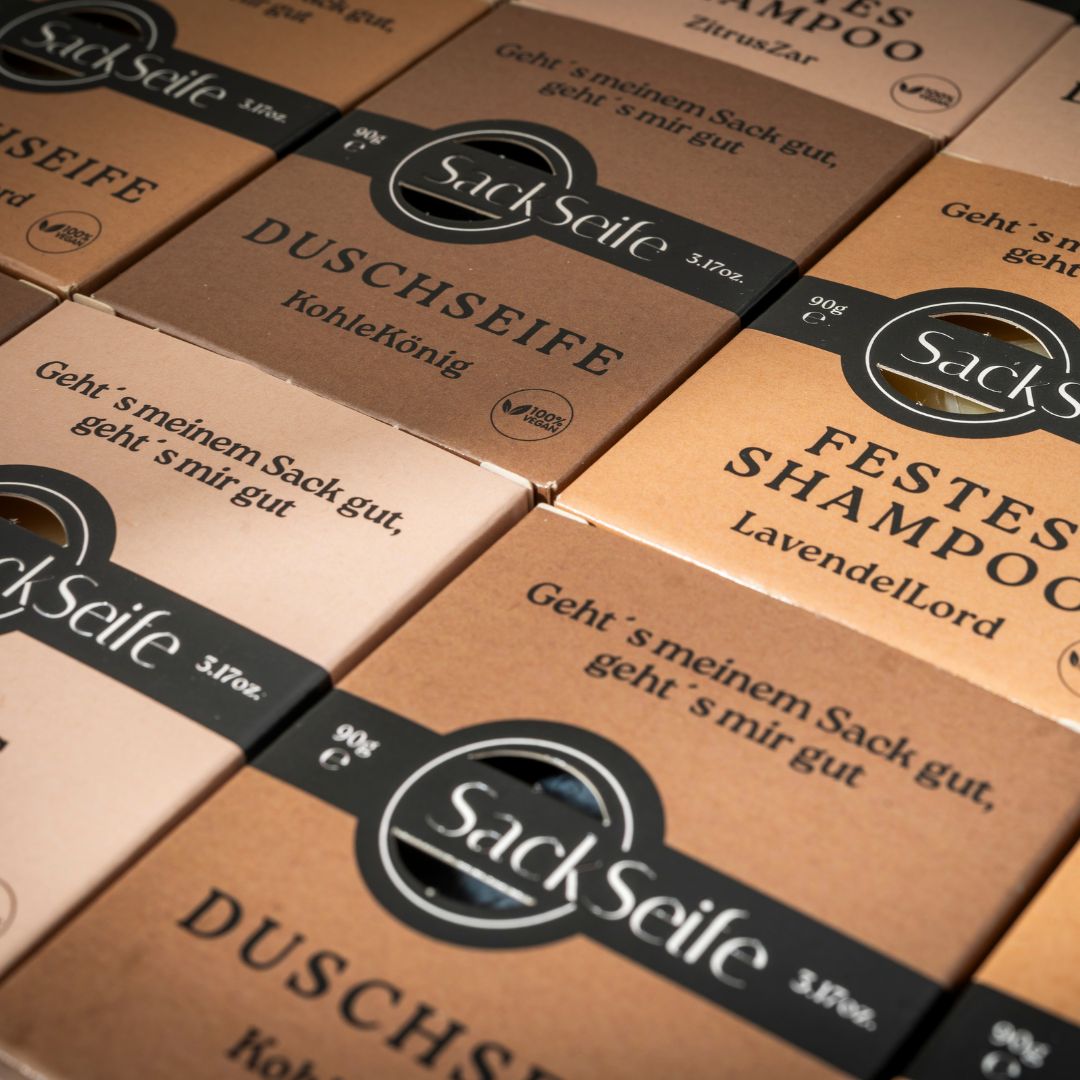 Nahaufnahme von mehreren Kartons. Aufschrift: SackSeife, DUSCHSEIFE, FESTES SHAMPOO. Verpackung aus braunem Karton.