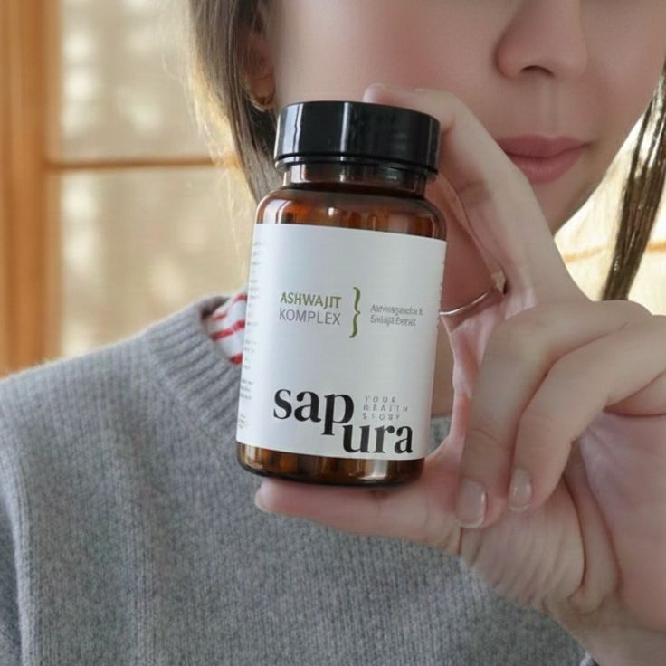 Braune Flasche mit schwarzem Deckel. Weißes Etikett mit Text: Ashwajit Komplex, Sapura Health. Frau hält Flasche.