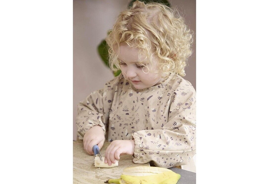Ein Kind mit Locken schneidet mit einem blauen Messer eine Banane auf einem Brett.
