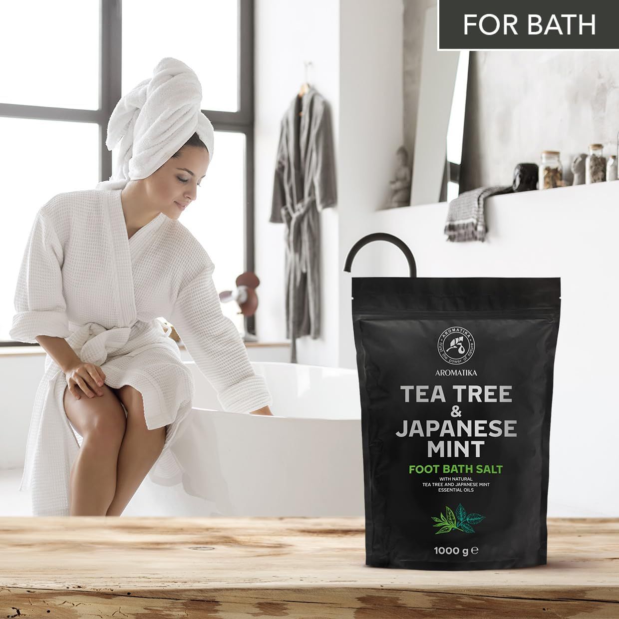 Frau im Bademantel neben Badewanne. Schwarze Verpackung mit Produktnamen und Logo. Text: Tea Tree & Japanese Mint Foot Bath Salt. 1000 g.