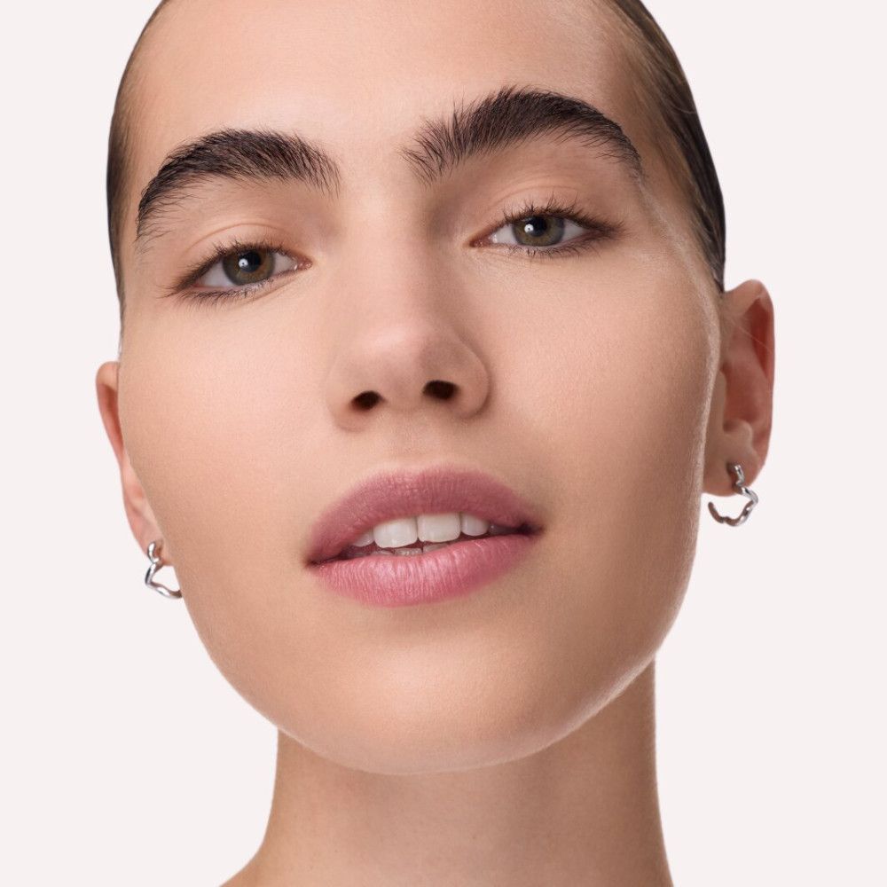Nahaufnahme eines Gesichts. Die Person hat helle Haut, dunkle Augenbrauen und trägt Ohrringe. Die Lippen sind leicht geschminkt.