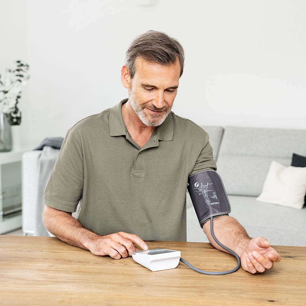 Mann benutzt ein Blutdruckmessgerät am Arm. Das Gerät liegt auf einem Tisch. Manschette am Oberarm. Graue Manschette.