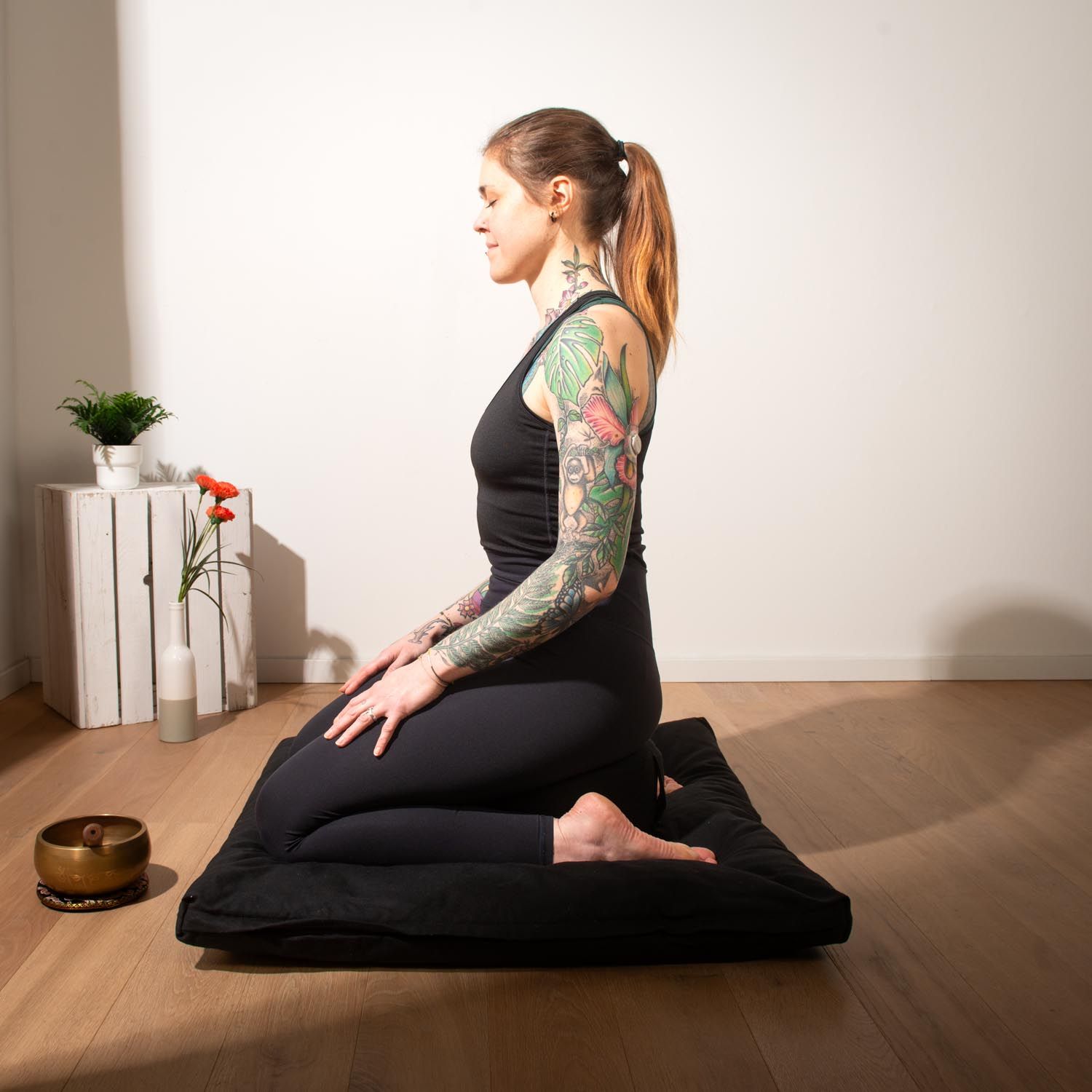 Person sitzt auf einem schwarzen Meditationskissen und Matte. Person trägt schwarze Kleidung.