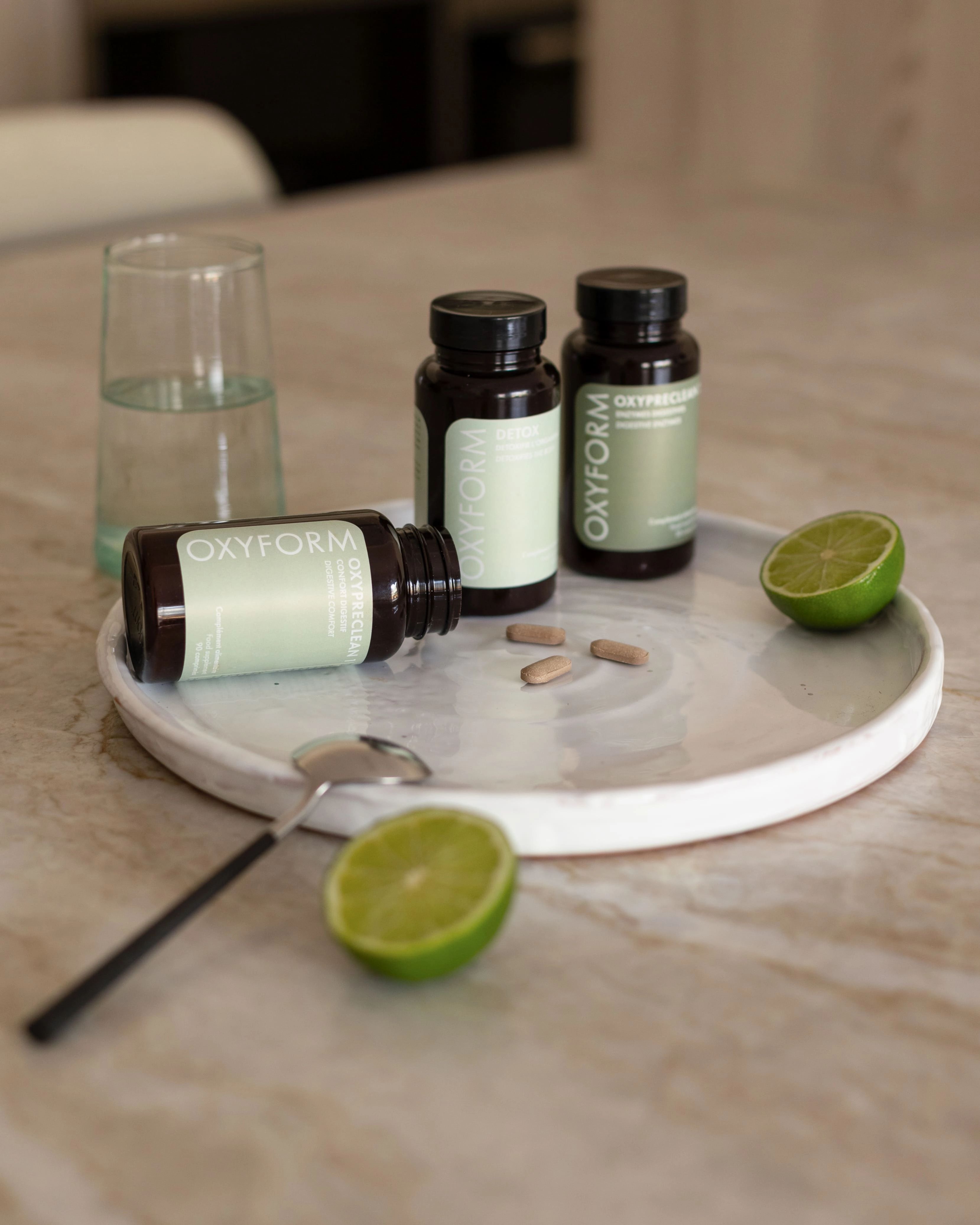 Drei braune Flaschen mit Etiketten, Tabletten, eine Limettenscheibe, ein Glas Wasser und ein Löffel auf einem Teller.